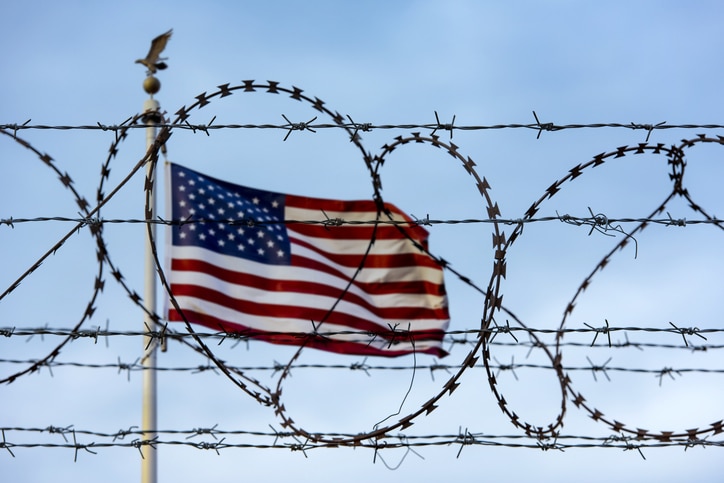American flag behind barbed-wire fencing representing detention